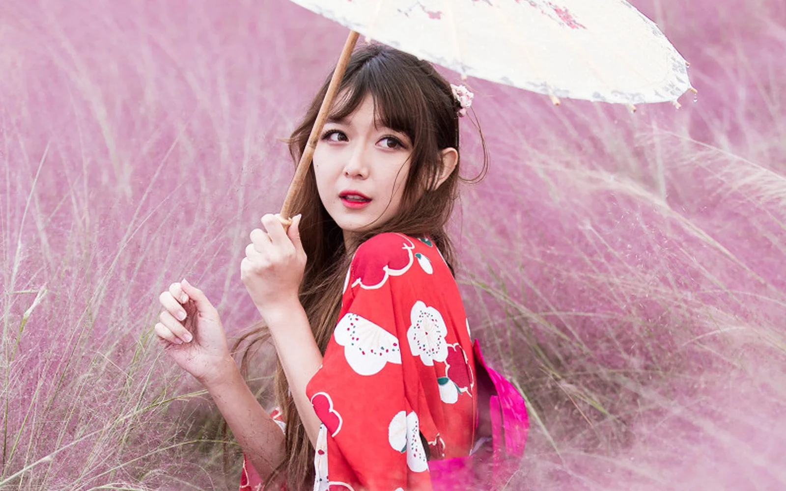 女性が白い傘を差し、ピンク色の草むらの中で赤い柄の服を着て立っている。