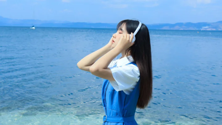 一名戴着耳机的女子站在清澈的海水旁，远处是蓝色海面与山脉。