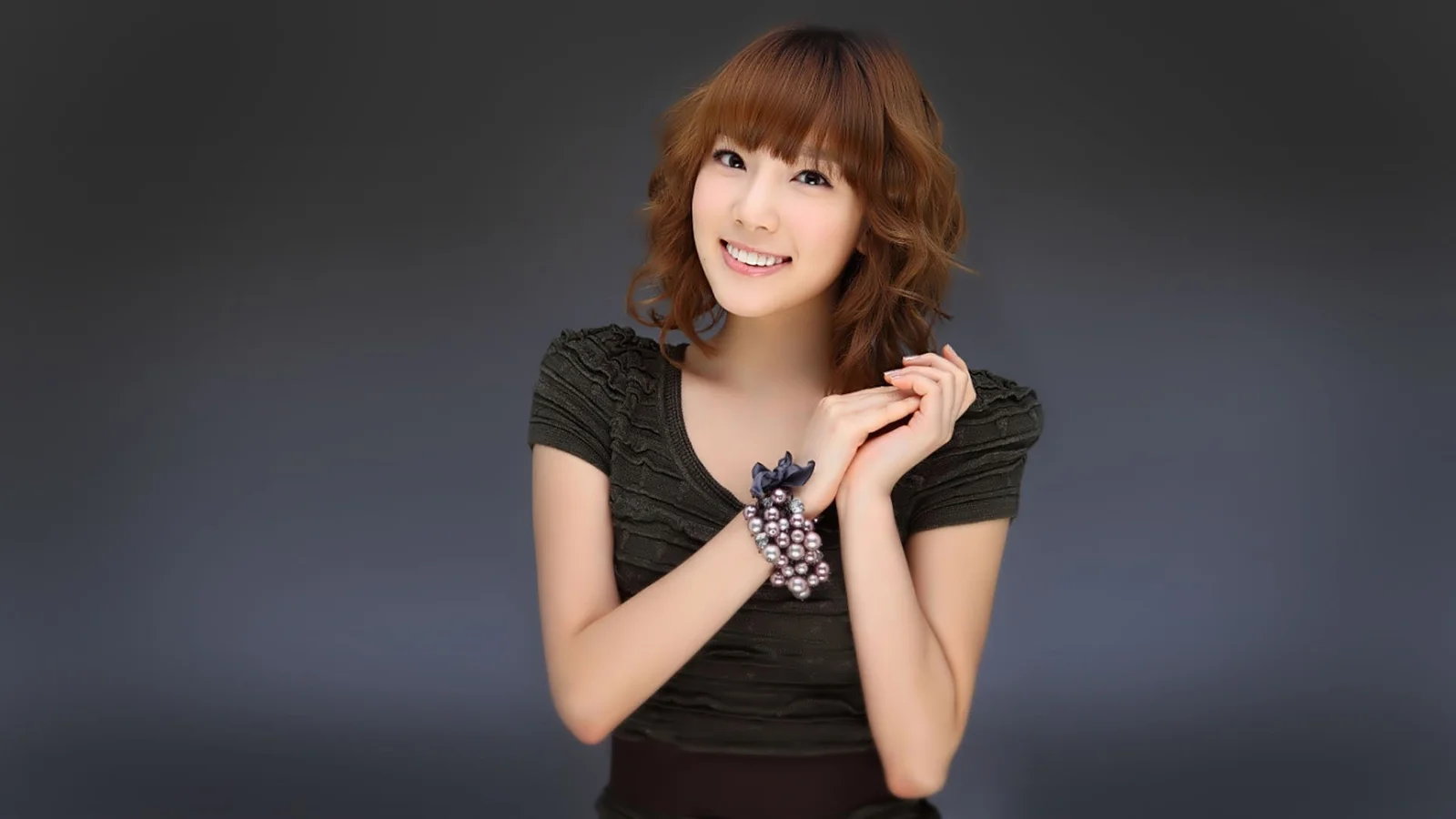 검은색 상의를 입은 성인 여성이 짙은 회색 배경 앞에서 미소를 지으며 포즈를 취하고 있습니다.