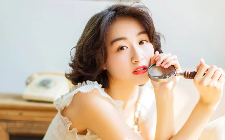 一名成年女性手持放大镜面对镜头微笑，背景为明亮的室内环境。