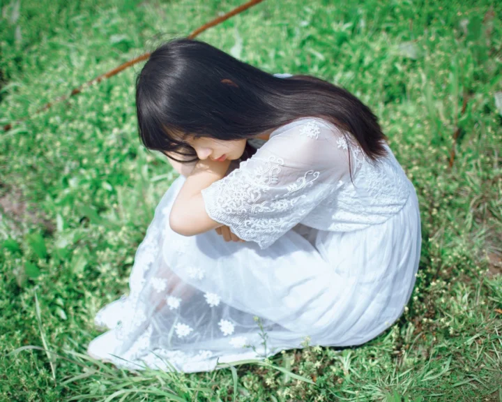 一名穿白色连衣裙的女子坐在绿草地上低头抱膝。