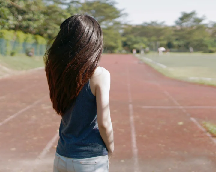 一名女性背对镜头站在户外跑道上，周围是树木和草地。