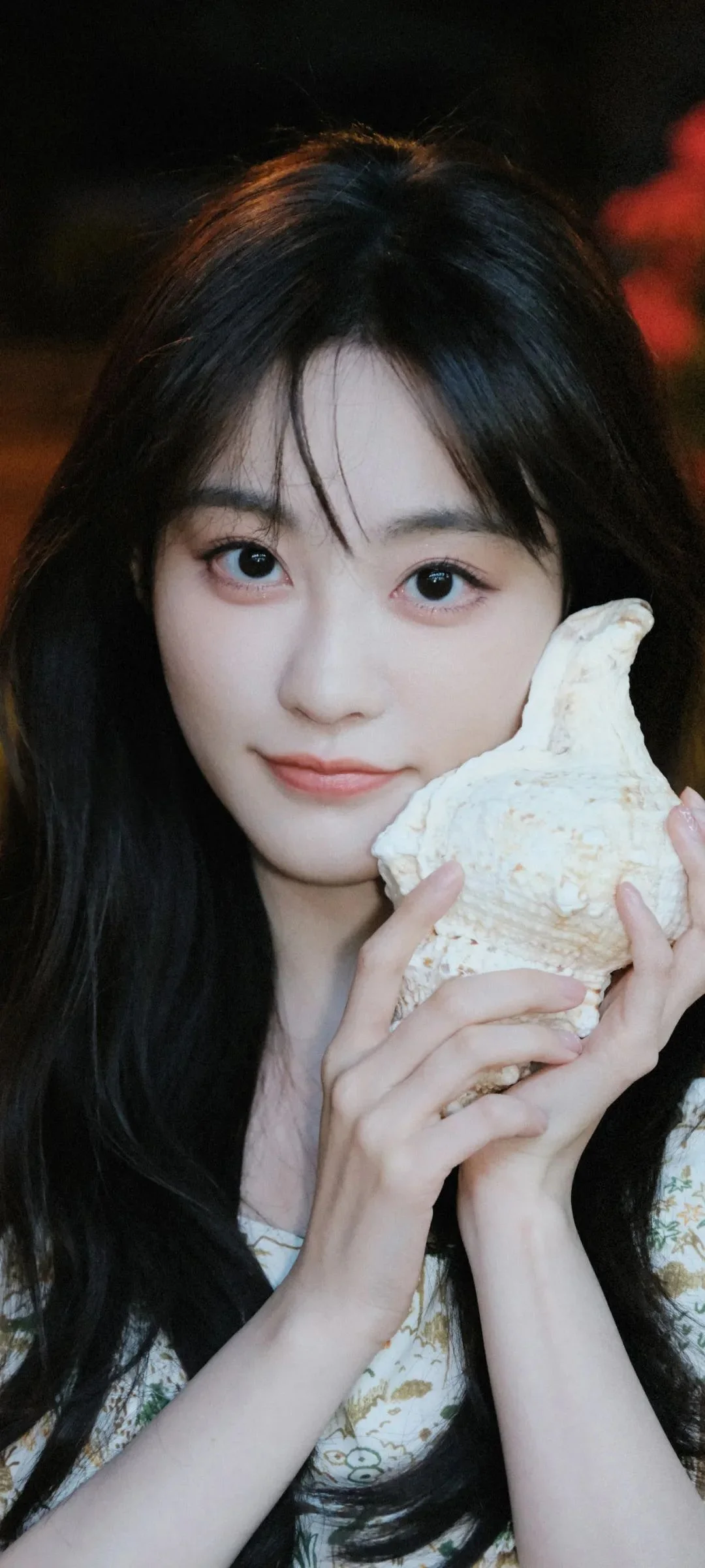 긴 머리의 여성이 양손으로 큰 조개껍데기를 들고 카메라를 바라보고 있습니다.
