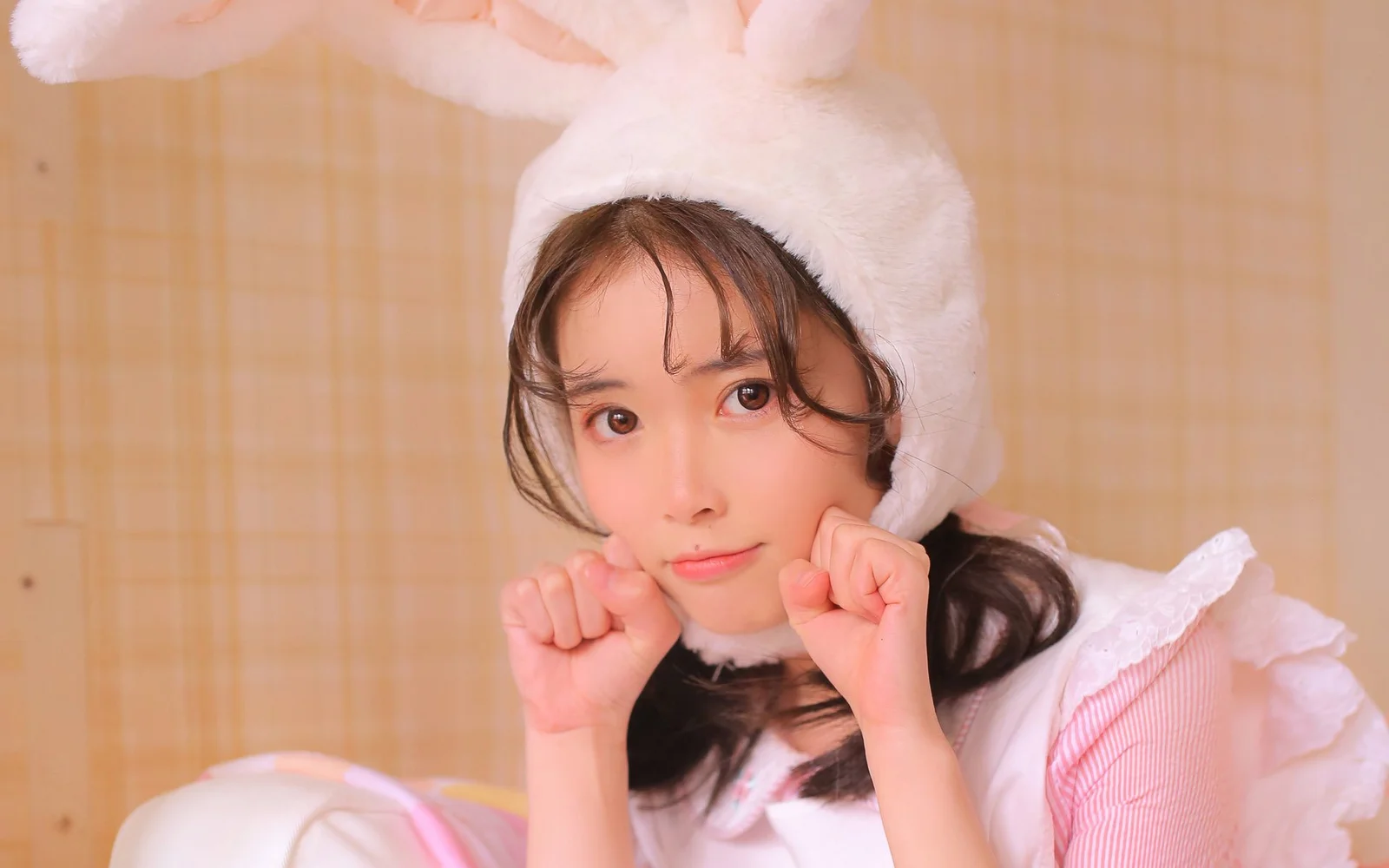 白いウサギ耳の帽子をかぶった女性が、暖色系の背景の前で頬杖をついて可愛らしくポーズを取っています。