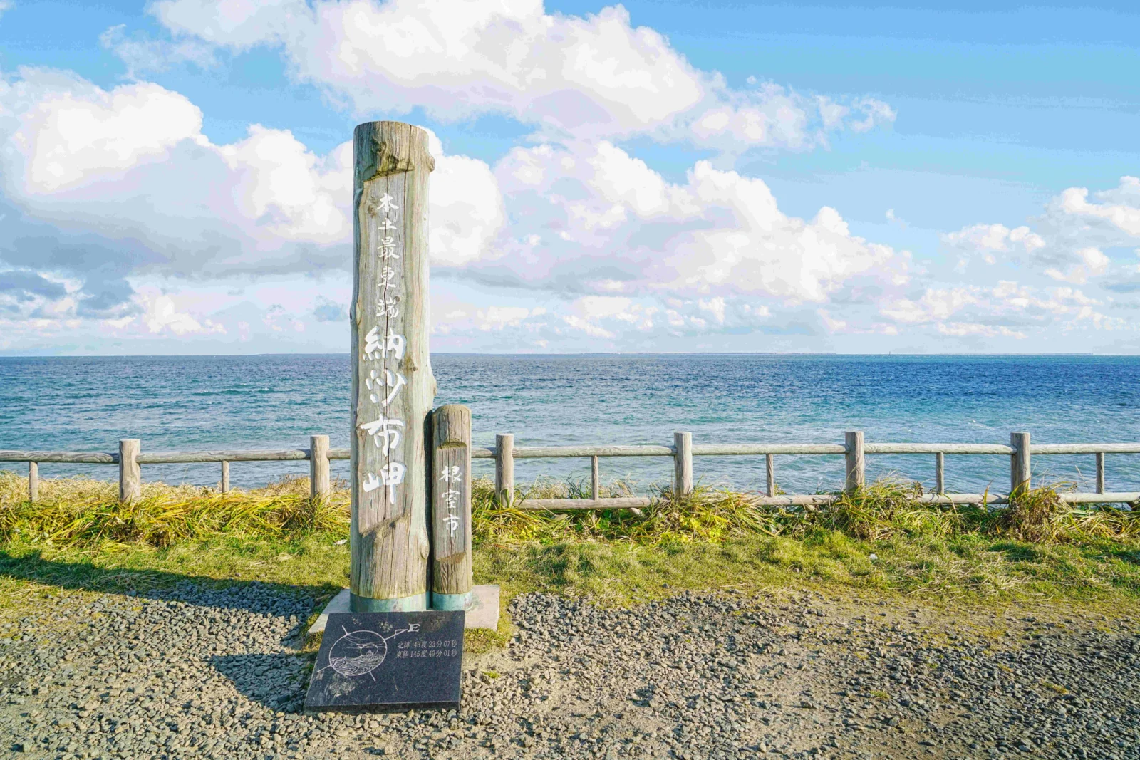 一塊木製標牌立在卵石海岸前，遠處是平靜的藍色海面和大片白雲。