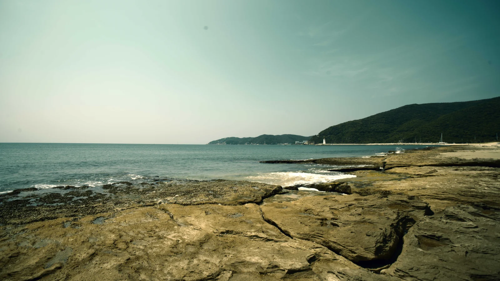 岩石海岸延伸至宁静海湾，远处是起伏的绿色山丘与开阔天空。