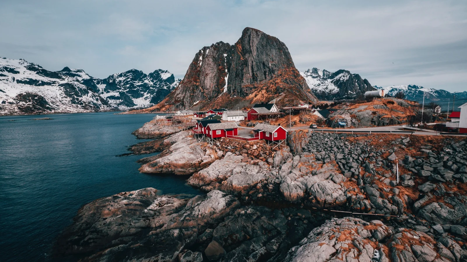 A Nordic coastal landscape with snow-covered mountains, a calm bay, rocky shores, and red waterfront cabins.