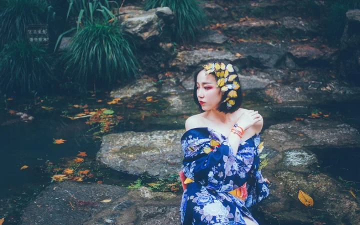 一名戴着花冠、穿蓝色花卉裙的女子坐在溪边岩石上。