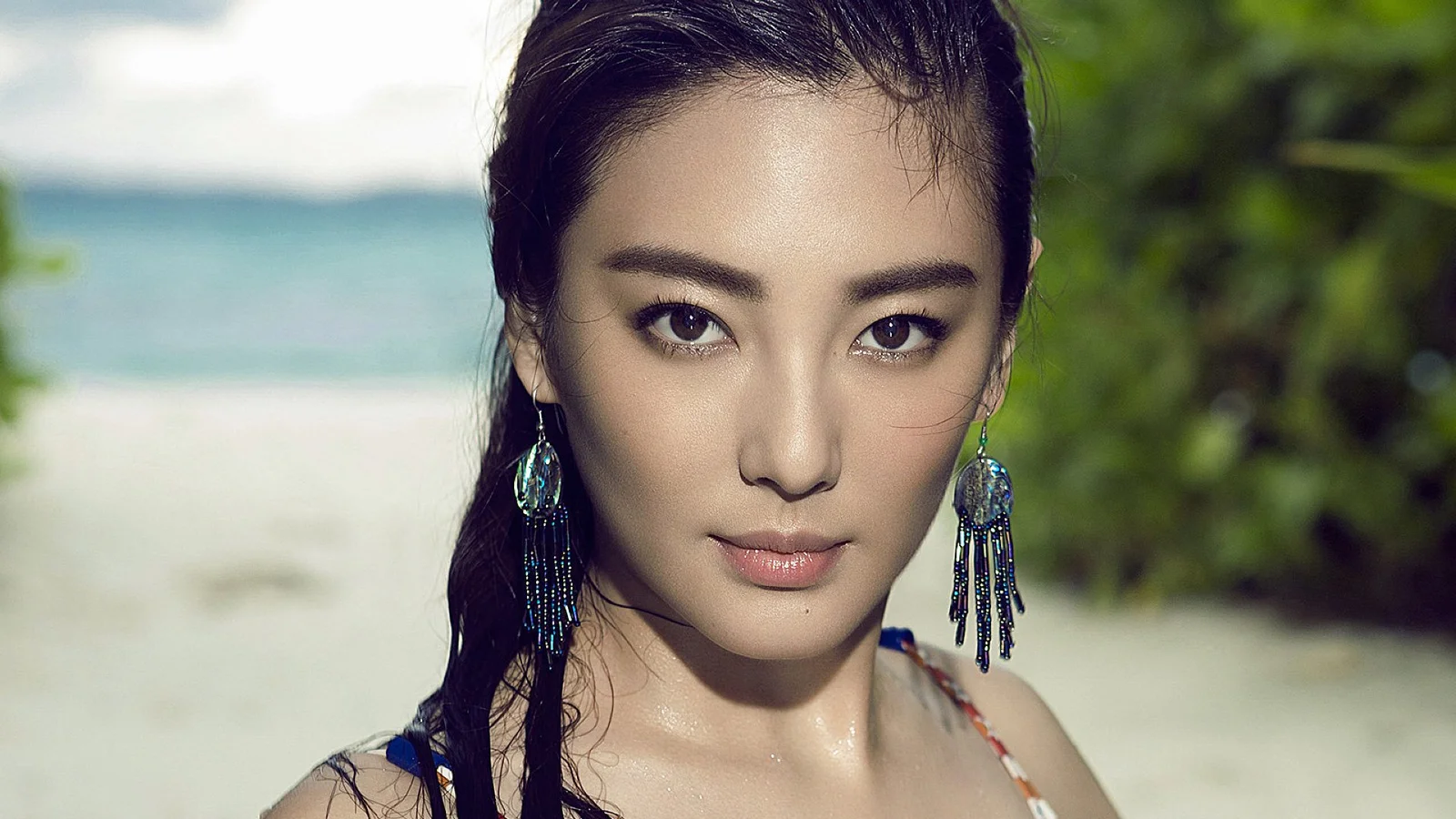 긴 귀걸이를 착용한 성인 여성이 해변을 배경으로 카메라를 바라보고 있습니다.