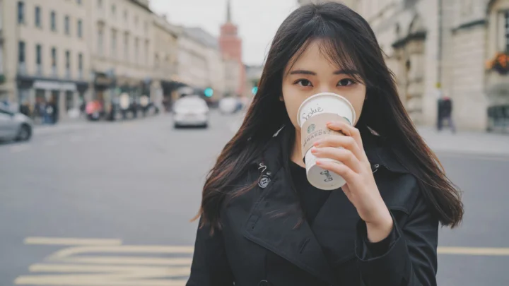 一名长发女子站在城市街道中央，手持咖啡杯遮住部分面庞。