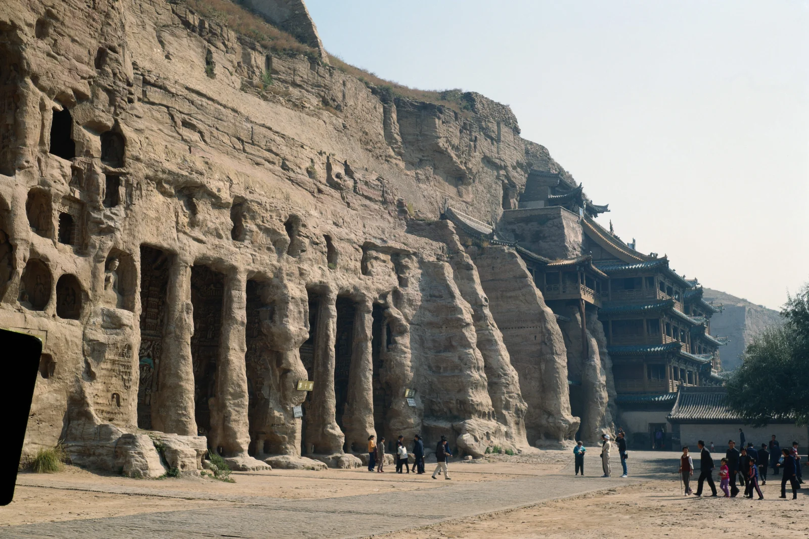 岩壁上的石窟与依山而建的古寺构成一幅户外历史遗迹景观，远处可见几名游客。