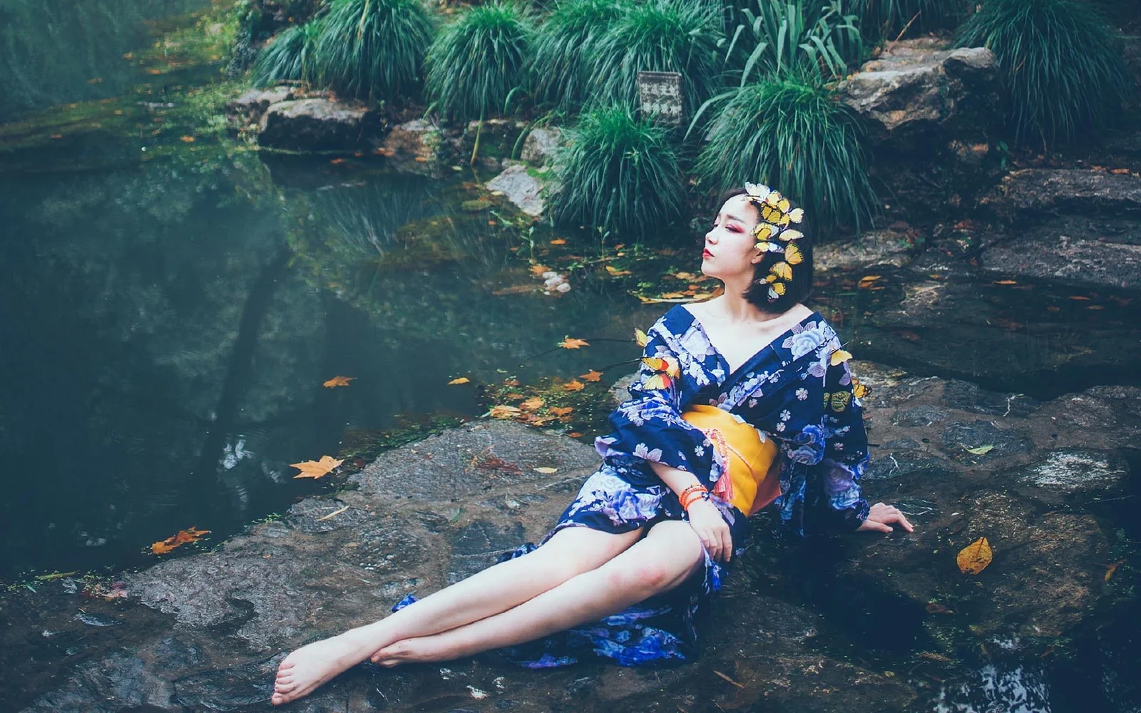 一名身穿传统服饰的成年女性坐在溪流旁的岩石上。