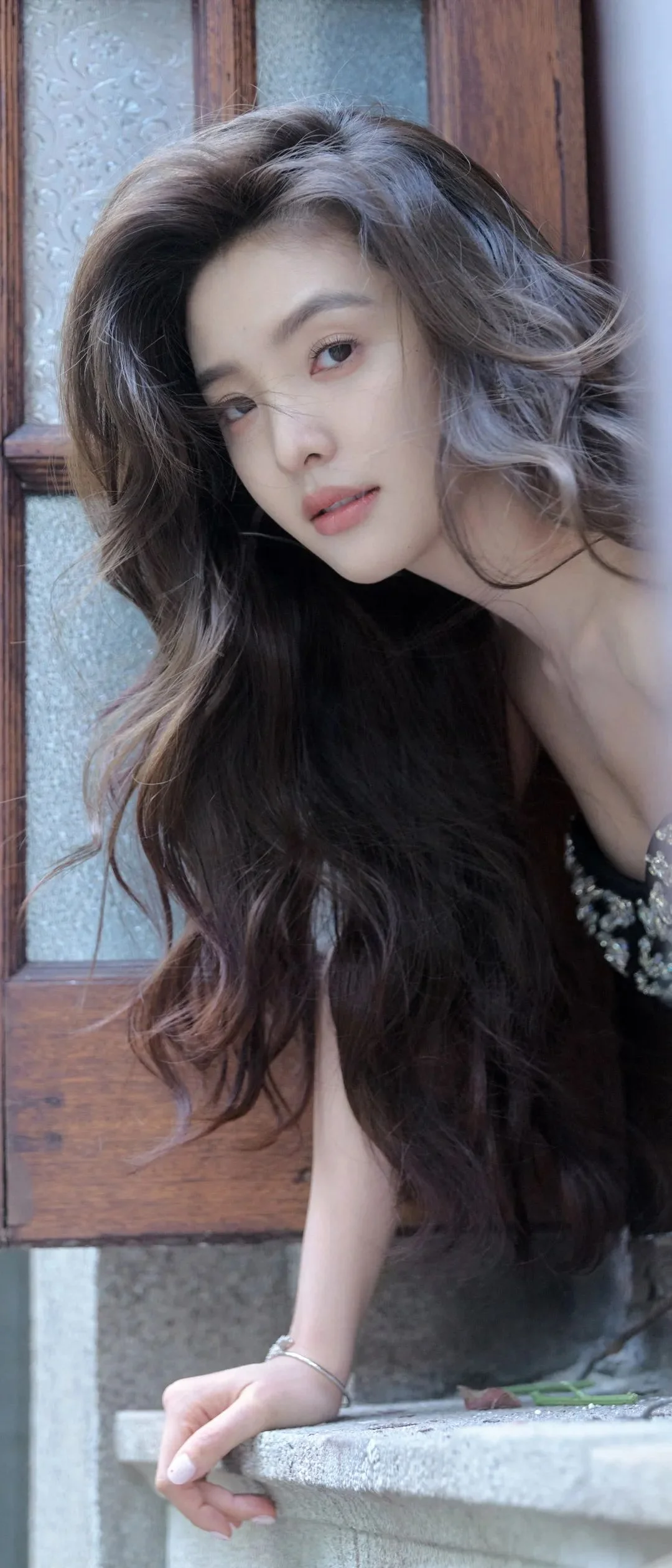 긴 머리의 여성이 창가에 기대어 부드러운 표정으로 카메라를 바라보고 있습니다.