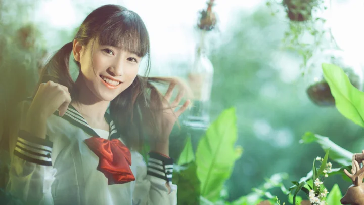 미소 짓는 여성이 푸른 잎으로 둘러싸인 밝은 야외에 서 있습니다.