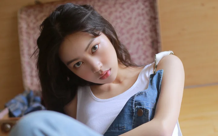 一名深色波浪长发女子穿着白色无袖上衣和牛仔背带裤，靠坐在木地板上。