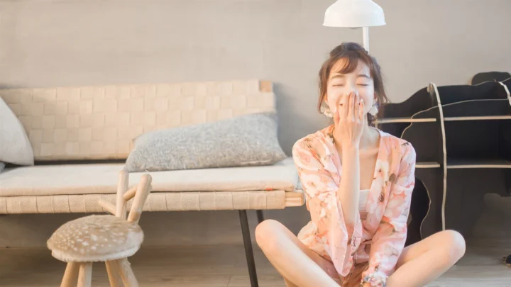 분홍색 꽃무늬 옷을 입은 성인 여성이 소파와 의자, 옷걸이가 있는 실내 바닥에 앉아 있습니다.