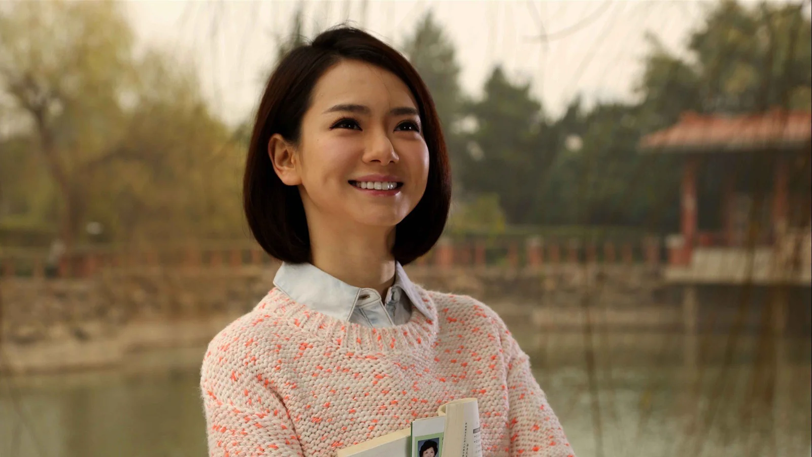 一位微笑的成年女性站在池塘边，手中拿着一本小册子。