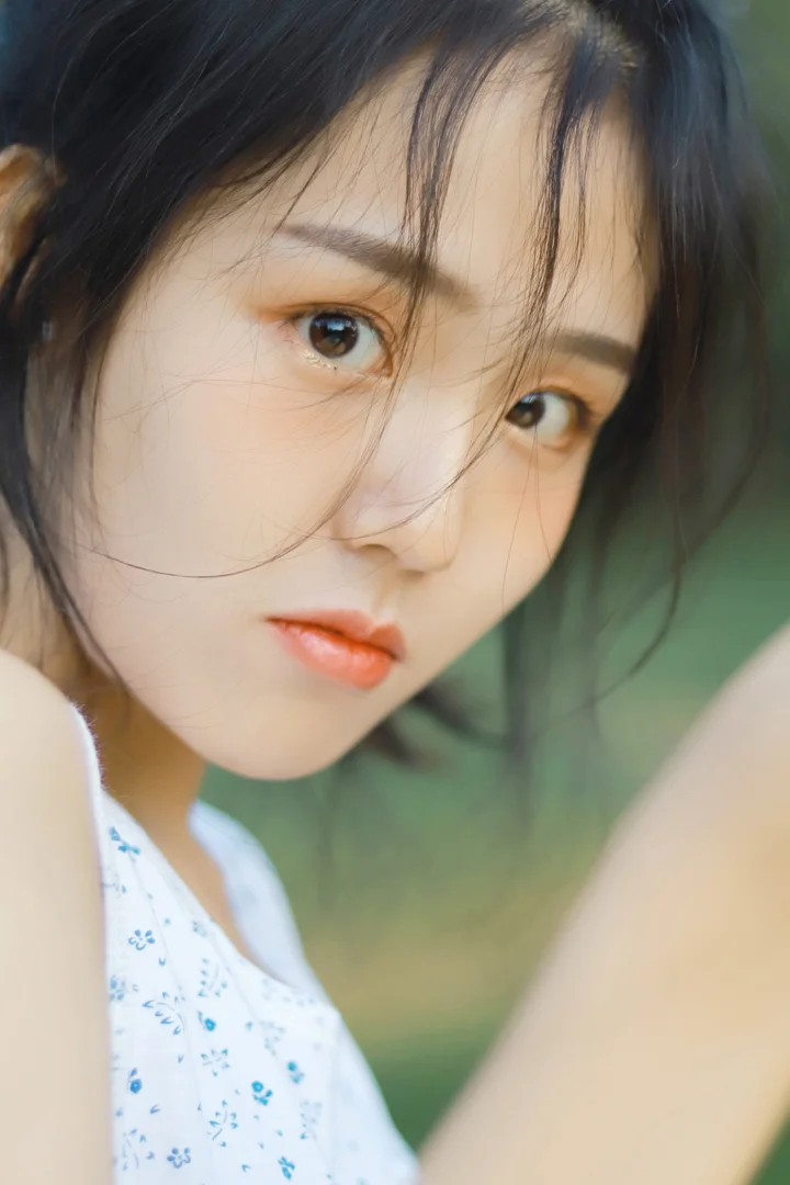 一名深色头发的女性近距离看向镜头，身穿白色蓝色碎花上衣，背景呈柔和的绿色虚化效果。