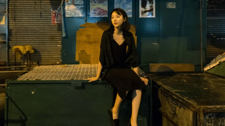 一名身穿黑色连衣裙的女子坐在昏暗城市环境中的金属平台上。