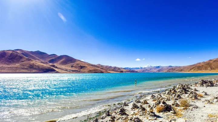 湛蓝湖水映衬着连绵群山，前景是阳光下的岩石湖岸。