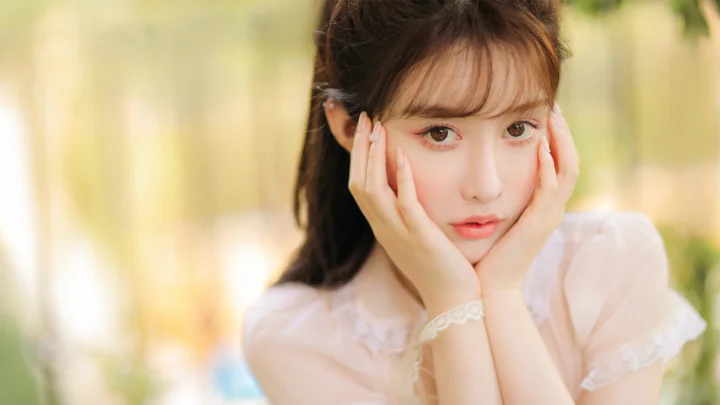 한 여성이 양손으로 볼을 받친 채 부드럽게 흐린 밝은 배경 앞에서 카메라를 바라보고 있습니다.