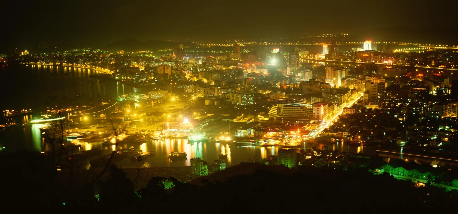 夜幕下的滨水城市灯火璀璨，建筑、道路与水面倒映出金色光芒。