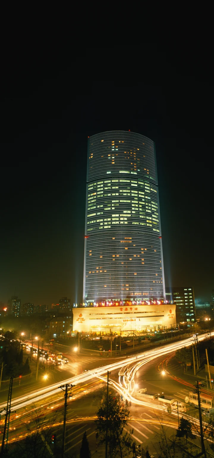 夜色中的高层圆柱形建筑矗立在繁忙路口上方，车流形成明亮的光轨。