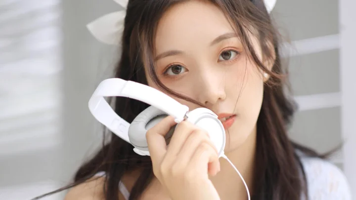 一名女性手持白色耳机面对镜头，背景明亮柔和。