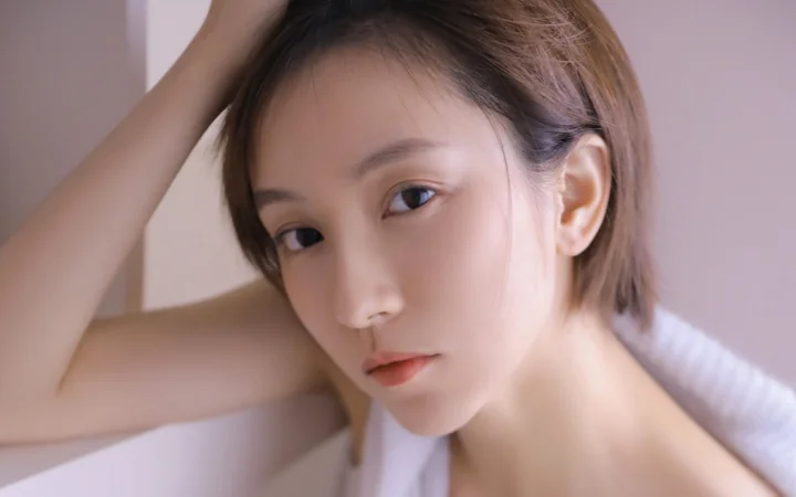 一位成年女性在柔和室内光线下靠近镜头摆姿势，手臂抬至头部旁边。