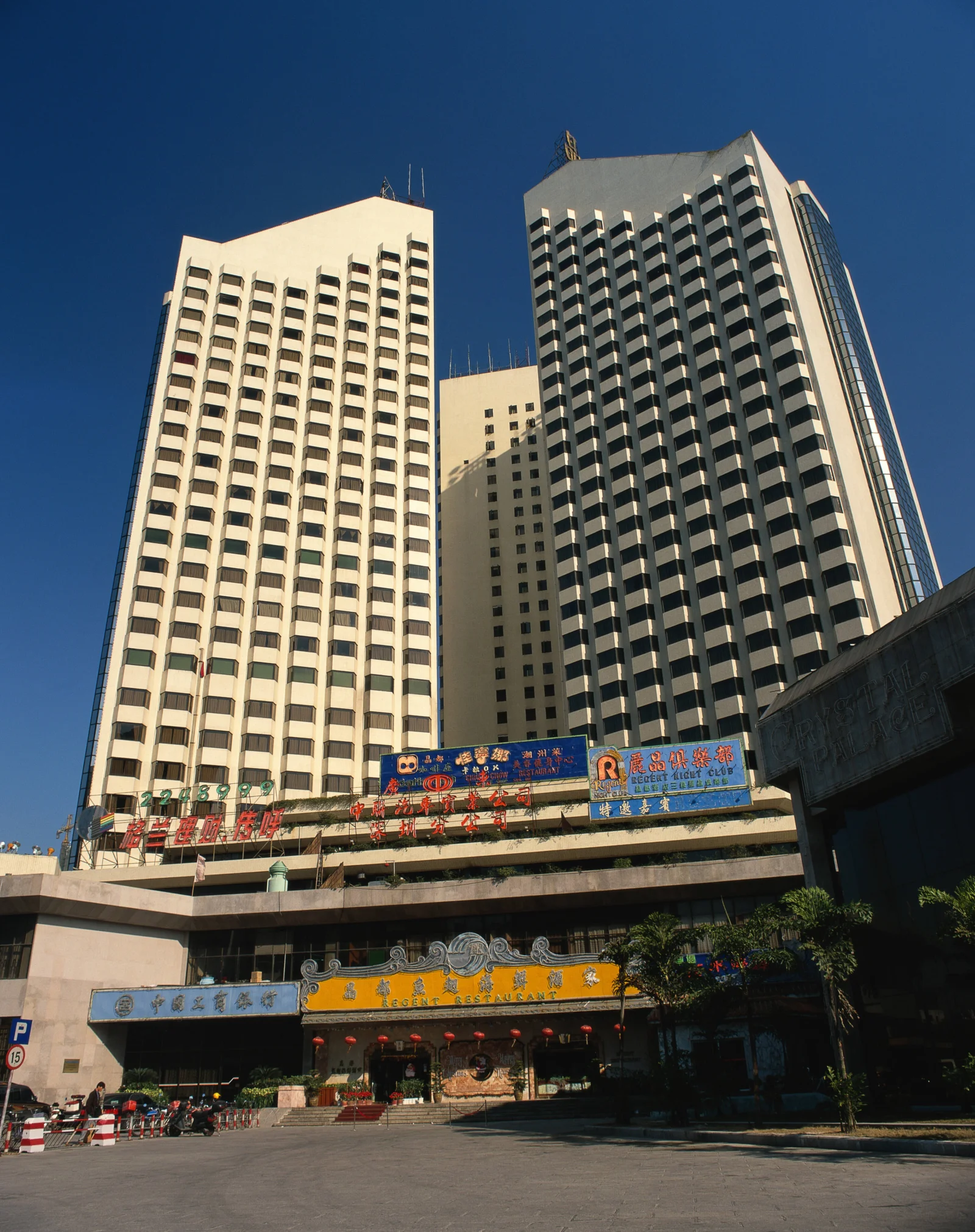 蓝天下，两座高层建筑矗立在城市街道旁，底部可见商铺招牌和入口。