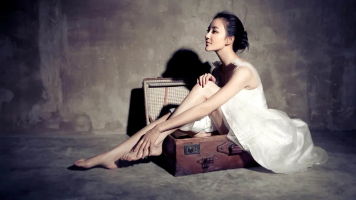 一名身穿白色连衣裙的成年女子坐在行李箱旁，置于昏暗的灰色墙面前。