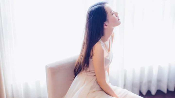 一名身穿白裙的女子坐在明亮窗边，柔和窗帘光线照亮了室内画面。