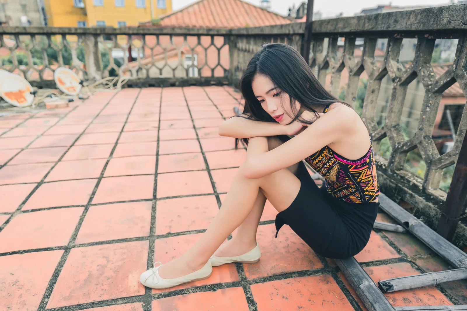 一名成年女子坐在铺着红色地砖的屋顶上，身后是栏杆和城市建筑。