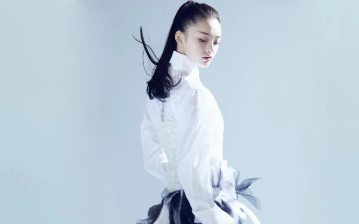 一名成年女性穿着白色高领上衣侧身站立，背景为浅蓝色纯色。