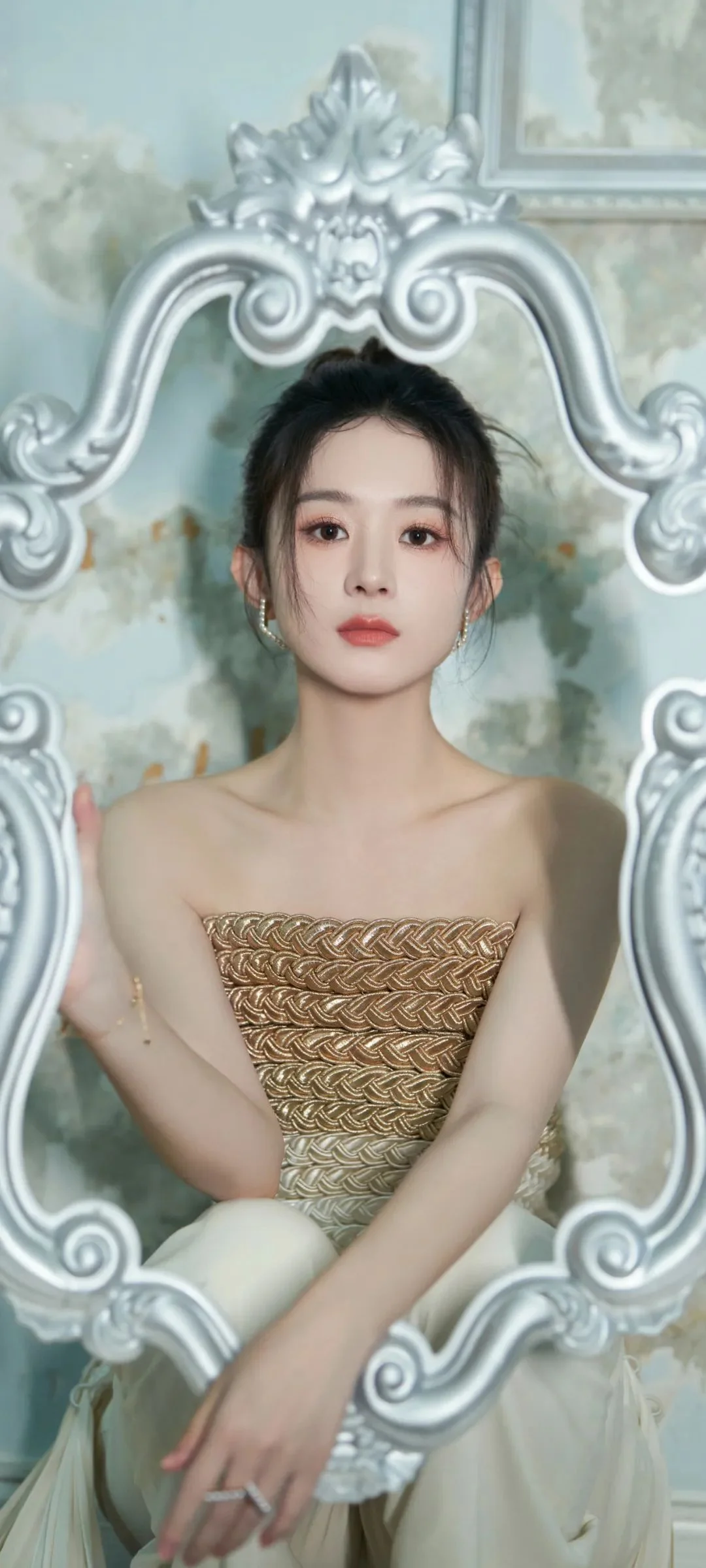 一名成年女性坐在华丽的银色装饰镜框前，穿着金色无肩带礼服。