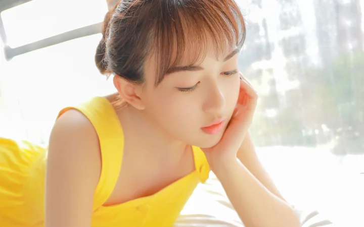 노란 민소매 상의를 입은 성인 여성이 밝은 창가에서 손으로 턱을 괴고 있다.