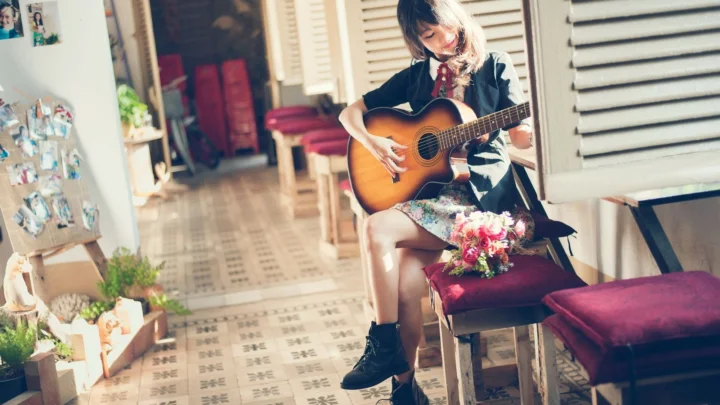 一名年轻女子坐在室内窗边弹奏木吉他，前景放着一束花。