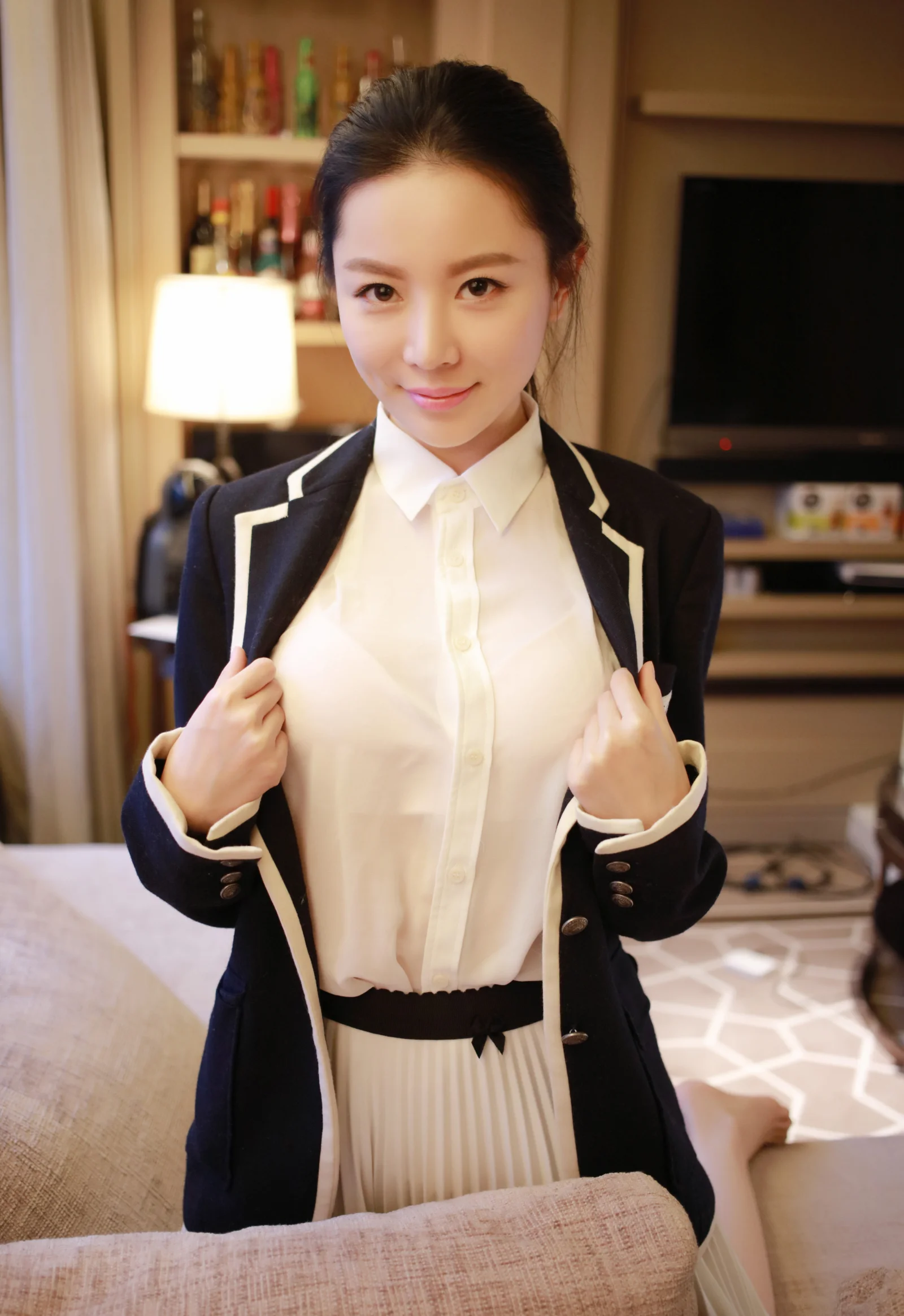 一名成年女性穿着深色外套和白色衬衫，在暖色灯光的室内面对镜头。