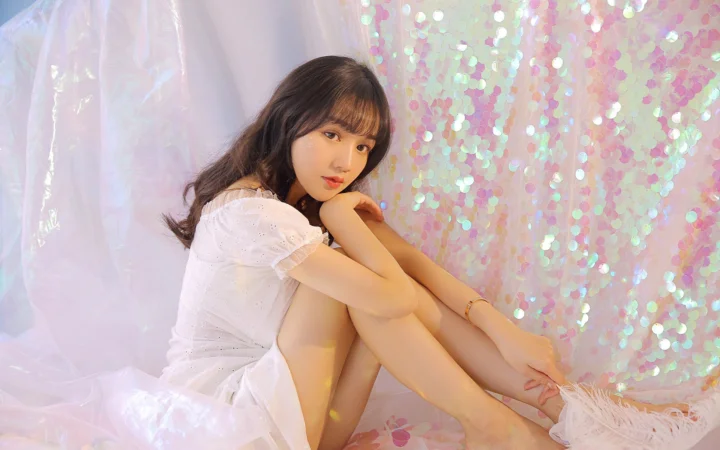 흰색 원피스를 입은 성인 여성이 부드러운 파스텔 색상의 스팽글 배경 앞에 앉아 있습니다.