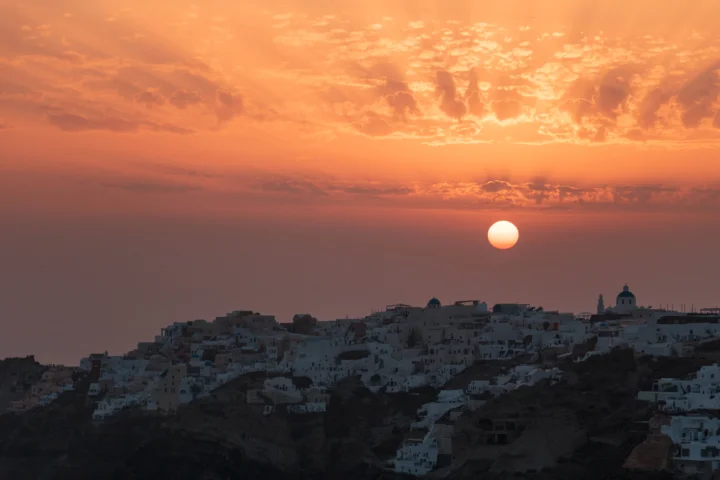 夕阳悬挂在圣托里尼白色山城上空，天空被染成橙红色。