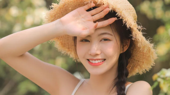 밀짚모자를 쓴 성인 여성이 야외에서 손으로 햇빛을 가리며 미소 짓고 있습니다.