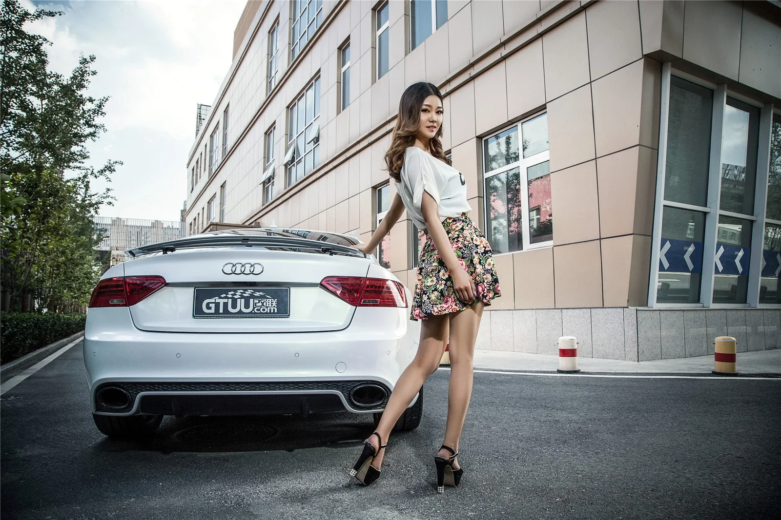 一名成年女性站在城市建筑旁的银色汽车后方。