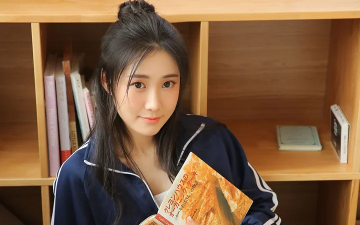長い髪の女性が濃い色のスポーツジャケットを着て、本棚の前で本を持っている。