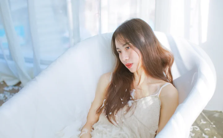 一名成年女子穿着白色连衣裙坐在白色扶手椅上，沐浴在明亮柔和的室内光线中。