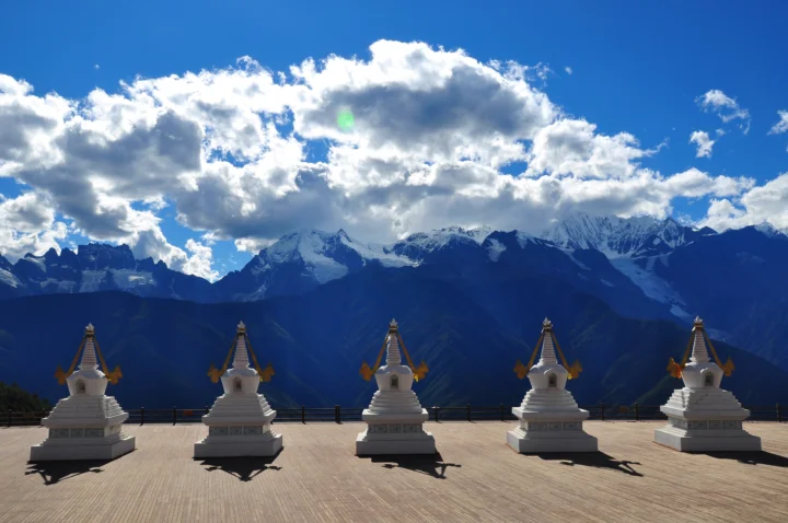 藍天白雲下的雪山前排列著幾座白色佛塔。