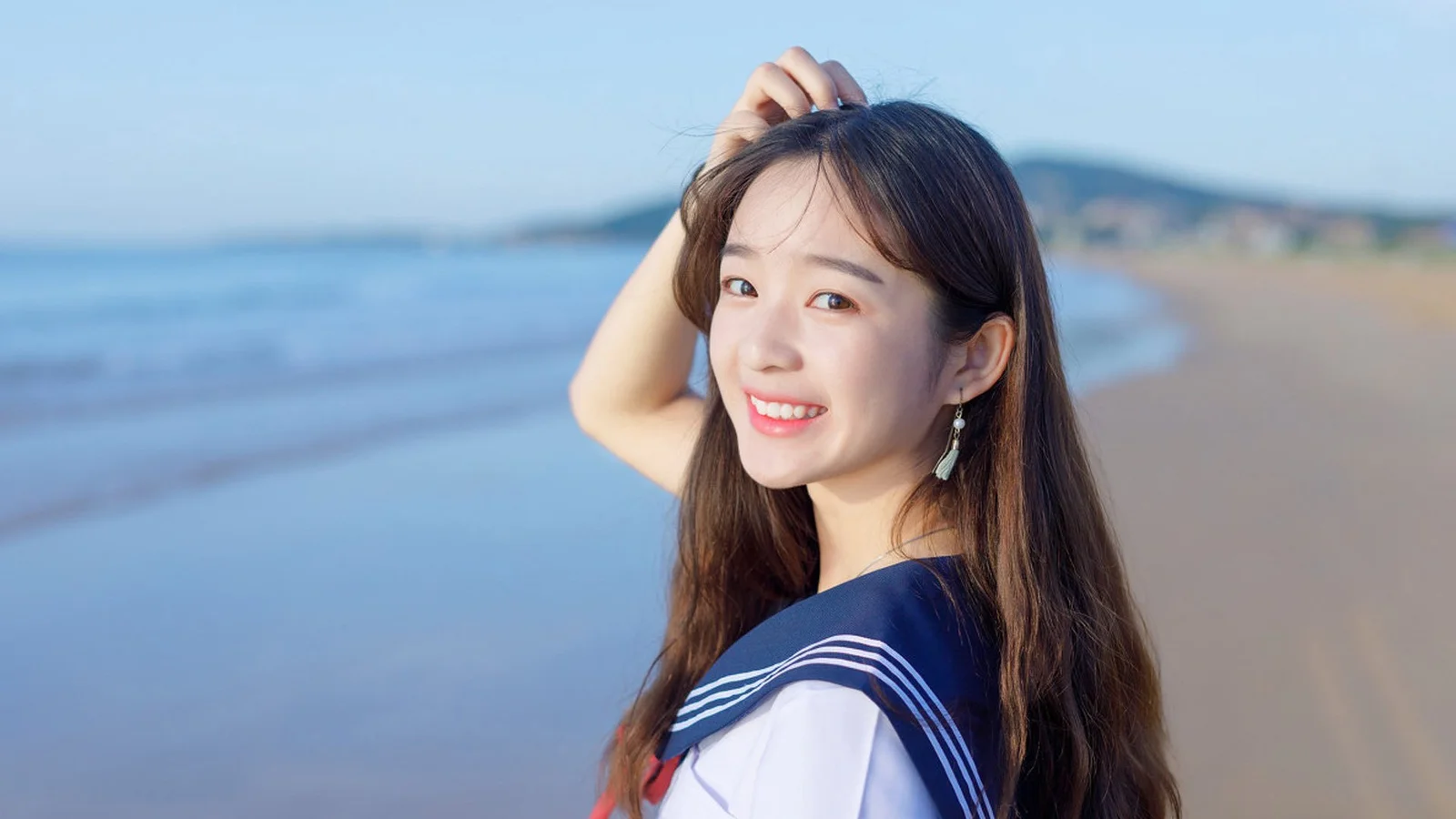 긴 머리의 여성이 해변에서 카메라를 향해 미소 짓고 있으며 뒤로 푸른 바다와 모래사장이 보입니다.