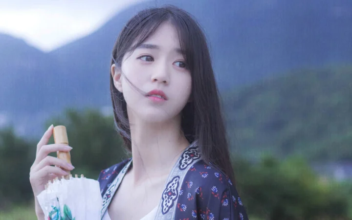 成人の女性が透明な傘を手に、山の風景を背景に立っている。