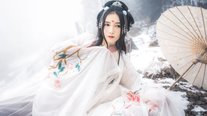 一名身穿白色传统服饰的成年女性坐在雪地中，身旁放着一把纸伞。