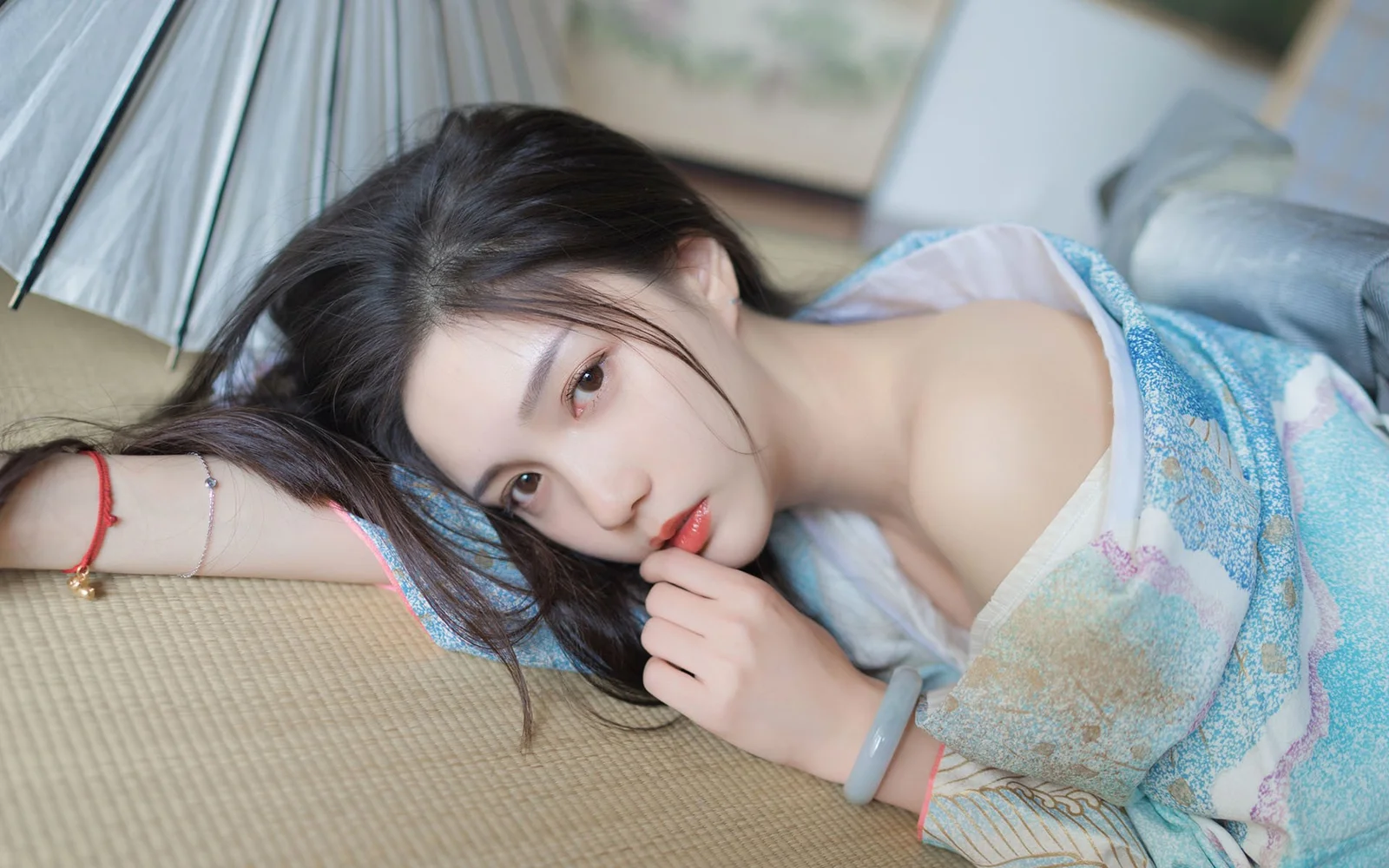 긴 머리의 여성이 짜임 매트 위에 누워 손가락을 입술 가까이에 대고 있으며 옆에는 밝은 색 우산과 흐릿한 실내 배경이 보인다.