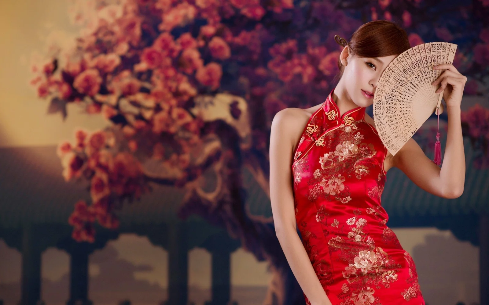 赤い花柄のドレスを着た成人女性が、花の背景の前で扇子を持っている。